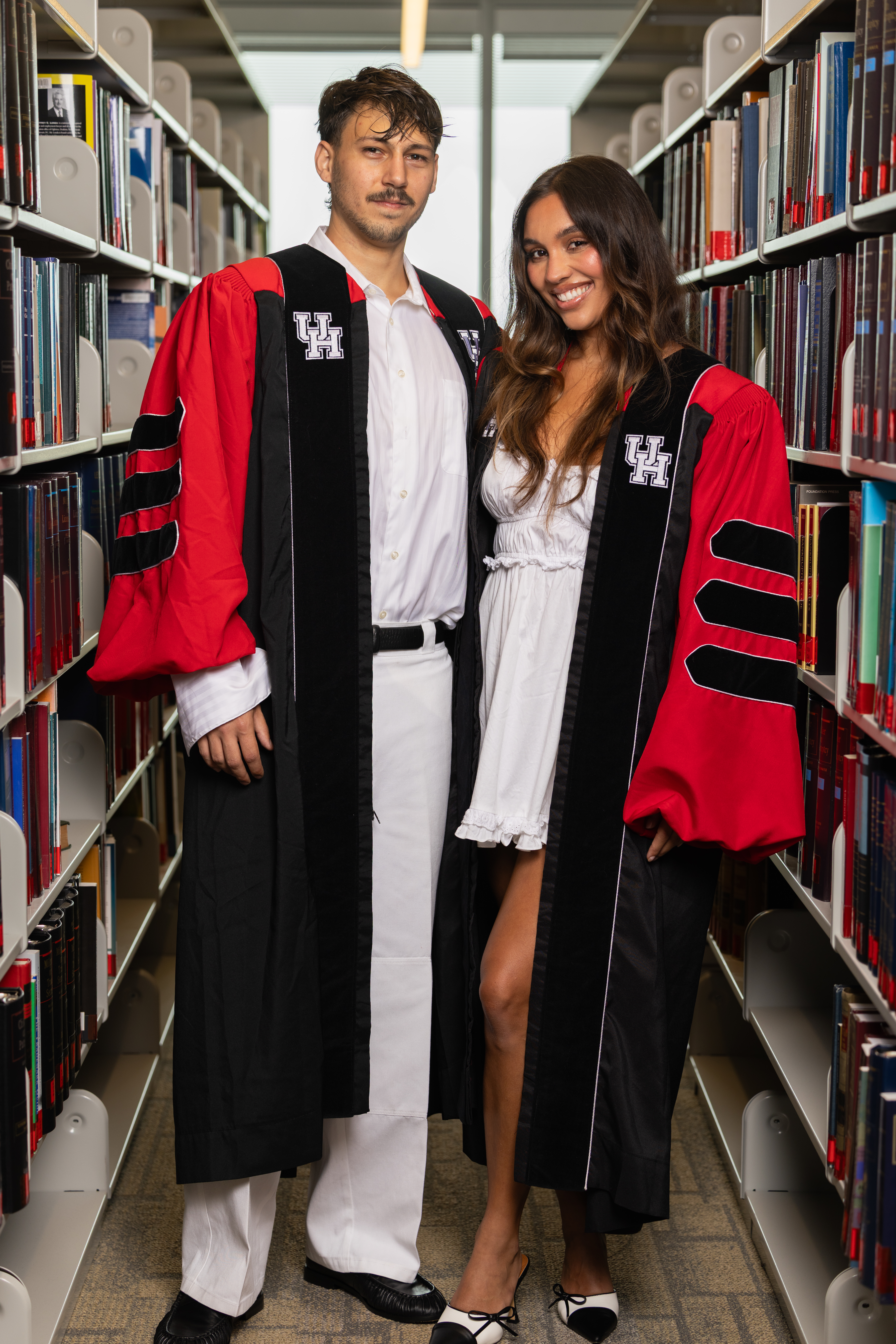 Emma Jenny and Tanner Shae stand together in graduation regalia in the UH Law Library.